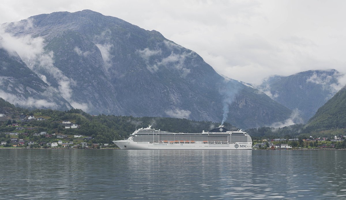 
Hvit cruiseskip i en fjord med skydekkede fjell i bakgrunnen.
