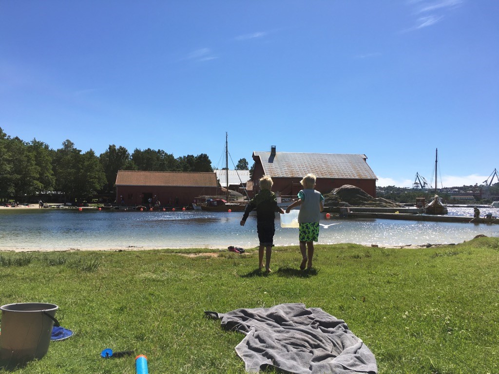 To gutter i niårsalderen går med et stort kar med vann i en liten vik utenfor Kristiansand. Solen glitrer i vannoverflaten, hvit sand og røde båthus i bakgrunnen 